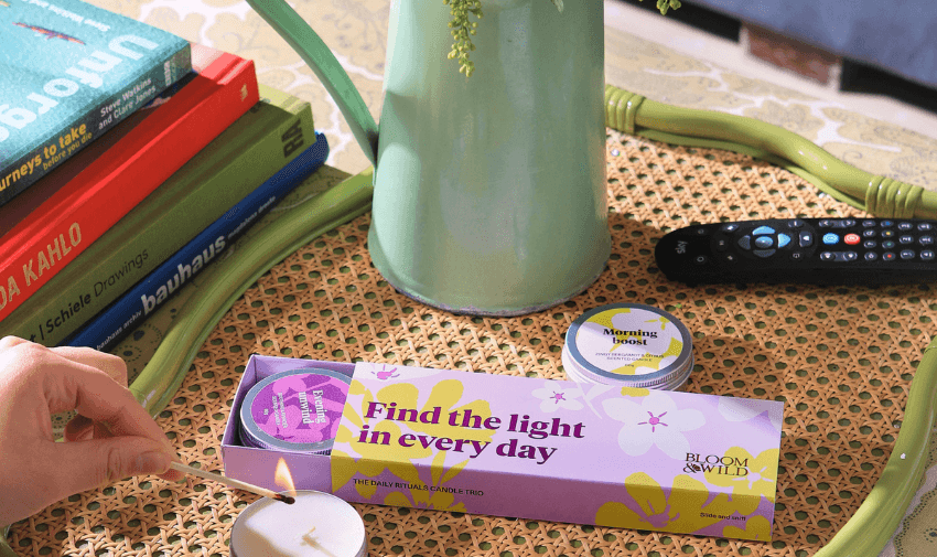 A hand lighting a candle next to a box of Bloom & Wild candles on a wicker tray, surrounded by books and a TV remote. The candle box reads "Find the light in every day," ideal for a relaxing home environment. Disabled discounts on wellbeing products.