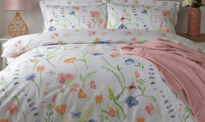 White bedding featuring a colourful floral and butterfly pattern. A pink knit blanket is draped over the edge of the bed.