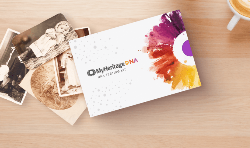 A MyHeritage DNA testing kit box is placed on a wooden surface with a vibrant, colourful design on the right. Nearby are vintage black-and-white photographs, and a cup of coffee is partially visible in the top right corner.