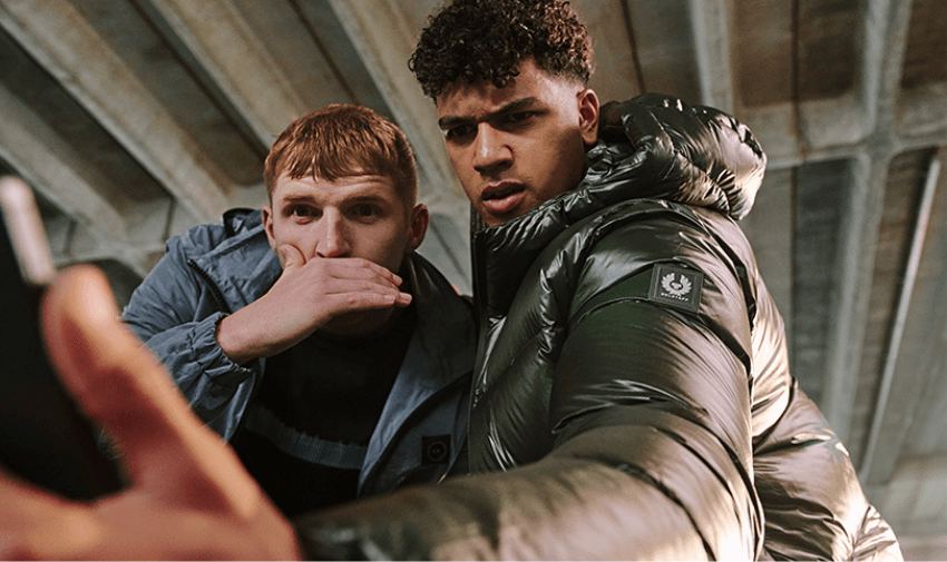 Two young men wearing stylish jackets, looking intently at a smartphone, with an urban setting of concrete beams in the background.