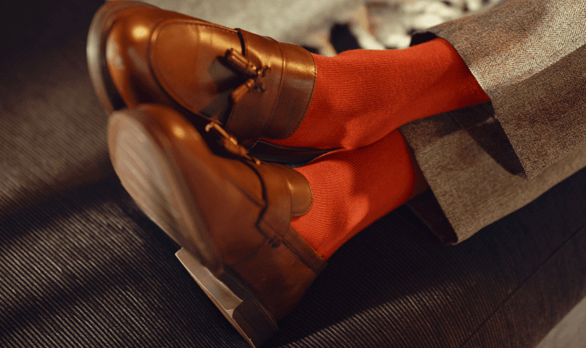 A pair of brown leather loafers with tassels is worn with bright red socks. The person is seated, with legs crossed, wearing grey trousers. The background is subtle and out of focus.