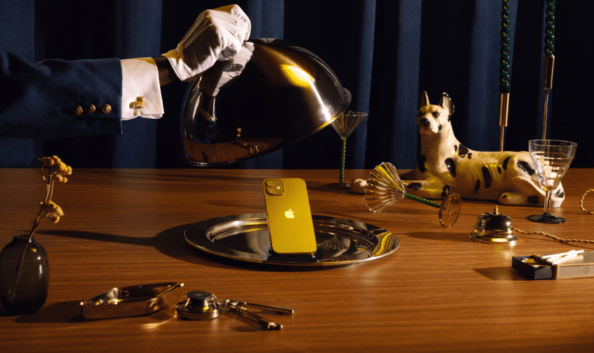 A white-gloved hand lifts a silver cloche to reveal a gold smartphone on a silver tray. The scene includes a porcelain dog figurine, green candles, a glass, and dried flowers, all set on a wooden table against a dark background.