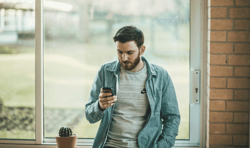 A man with short hair and a beard is casually leaning against a window ledge, looking at his smartphone. He is wearing a light denim shirt over a grey t-shirt. A small potted cactus is on the windowsill next to him.
