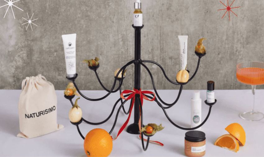 A black chandelier display features skincare products and small oranges on a table. A bag labeled "Naturisimo" is on the left, and a cocktail glass with an orange drink is on the right. A red ribbon is tied around the chandelier.