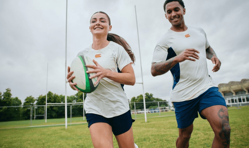 Two people are playing rugby on a grassy field. The person in front is holding a rugby ball, smiling, and running towards the camera. Both are wearing matching sportswear. Rugby posts and trees are visible in the background.