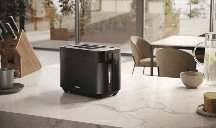 A sleek Haier Series 5 toaster in black sits on a marble kitchen counter. In the background, a knife block with knives, a cup, and a jug of water with lemon and mint are visible