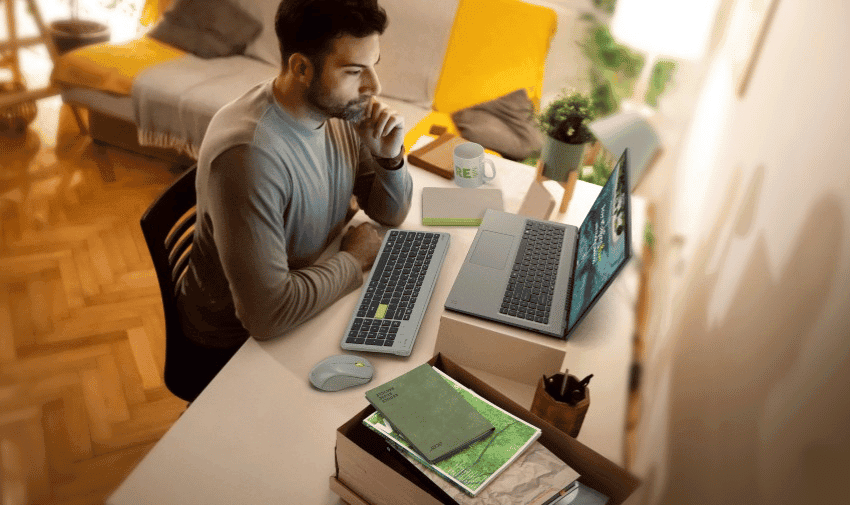 A man sits at a desk in a cosy, well-lit room, focused on the laptop in front of him. The desk also holds a keyboard, computer mouse and a stack of books.