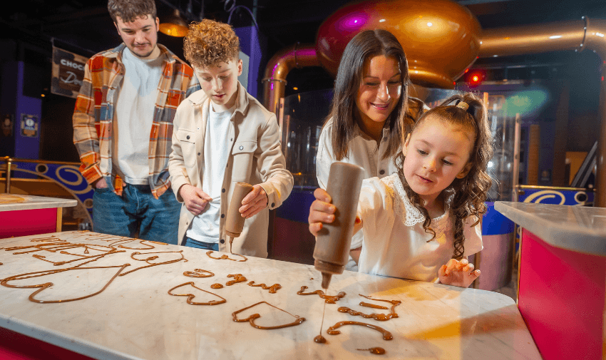 A family enjoys a hands-on chocolate-writing activity at Cadbury World, with children carefully piping words in chocolate onto a marble surface. A fun and educational experience for chocolate lovers. | Disabled attraction discounts, Cadbury World ticket offers.