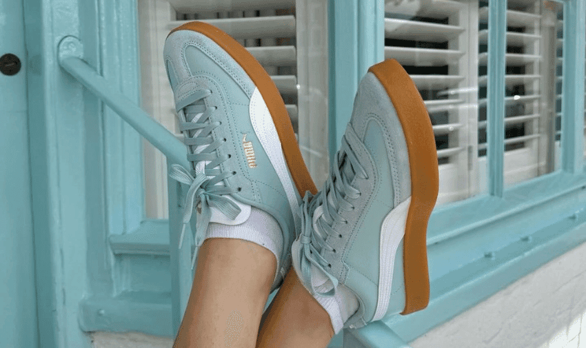 A person wearing light blue and white Puma sneakers with gum soles and light blue shoelaces is resting their feet against a light blue window frame with white blinds. They're also wearing white socks.