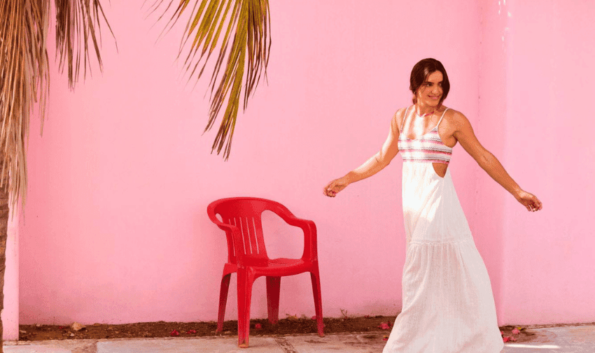 A woman in a white dress with colorful accents stands smiling against a pink wall. Her right arm is raised slightly, and a red plastic chair is positioned next to her on a concrete surface.