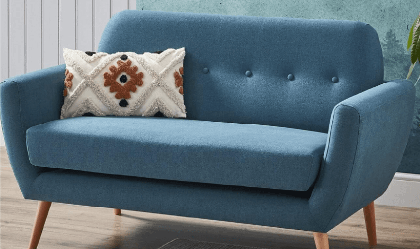 A mid-century modern blue loveseat with wooden legs is against a blue and white wall. It has a button-tufted backrest and is adorned with a textured white throw pillow featuring a diamond pattern in black and brown. The loveseat is placed on a wooden floor.