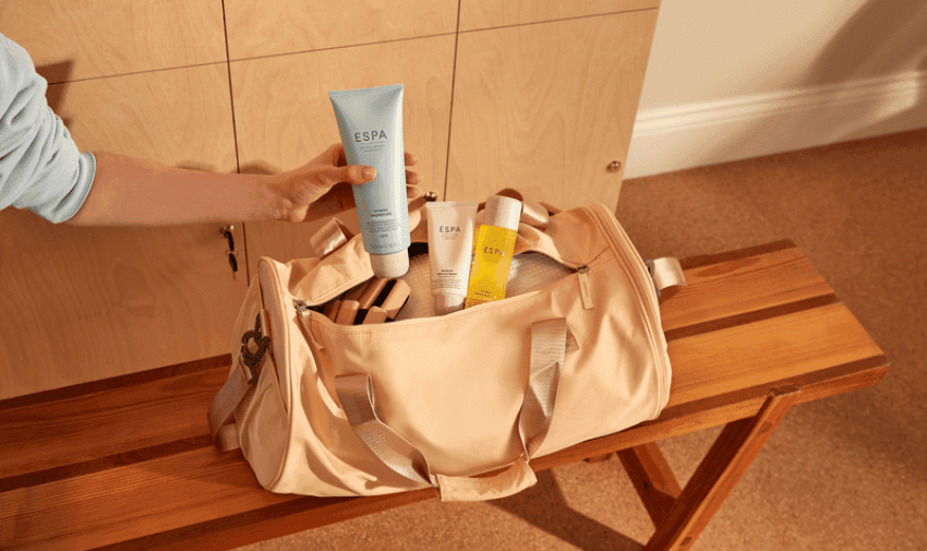 A hand reaching into an open beige gym bag on a wooden bench, holding a tube of ESPA product. Inside the bag, two additional ESPA product tubes are visible, one white and one yellow, against a light wood locker background.