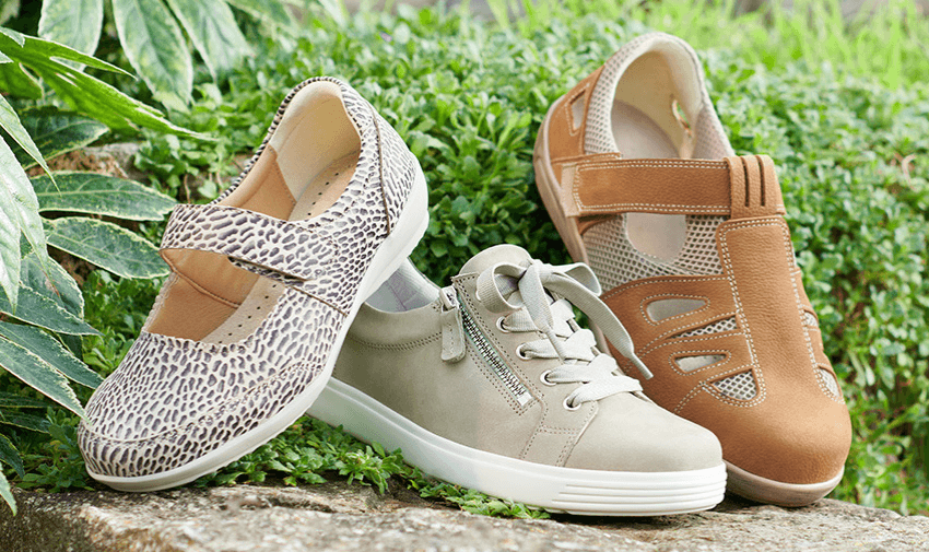 Three pairs of Cosyfeet wide-fitting shoes displayed outdoors on a stone ledge surrounded by greenery. Styles include beige trainers with a side zip, a tan velcro strap shoe, and a patterned Mary Jane style shoe. Mobility Discounts and Offers.