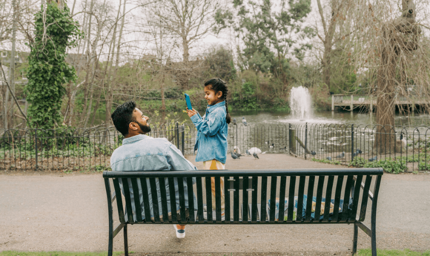 A person sits on a bench smiling at a child holding a phone in a park. The child stands on the bench, pointing the phone at the person. Trees, pigeons, and a fountain are visible in the background.