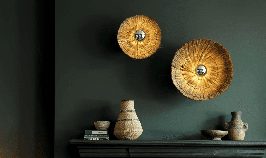 A dark green wall features two circular, woven straw wall lamps with glowing lights at their centers. Below, a black mantel holds a variety of decorative items including ceramic vases, a woven basket, and a stack of books. The scene has a modern, rustic aesthetic.