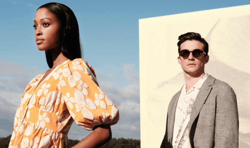 A woman in an orange floral dress and a man in a gray blazer with sunglasses stand outdoors against a sky backdrop and a large white screen. The woman faces forward, while the man looks slightly to the side. Both individuals appear confident and stylish.