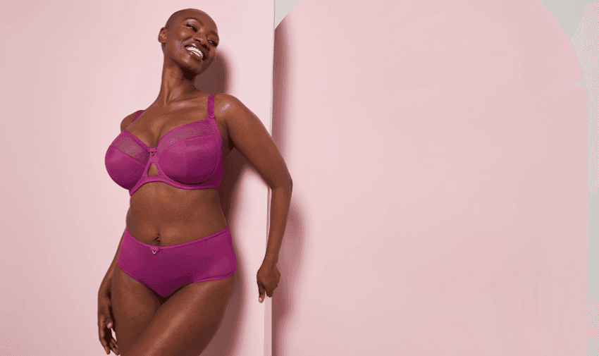 A woman is smiling and posing against a pink background. She is wearing a matching set of vibrant magenta lingerie.