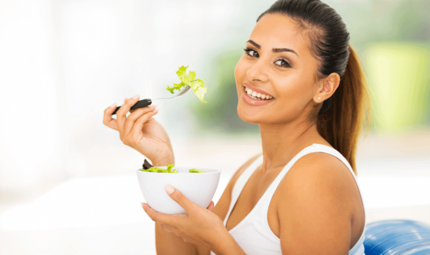 a lady is smiling at the camera, she is holding a white bowl filled with salad in her left hand, in her right hand she is holding a fork with lettuce leaves on