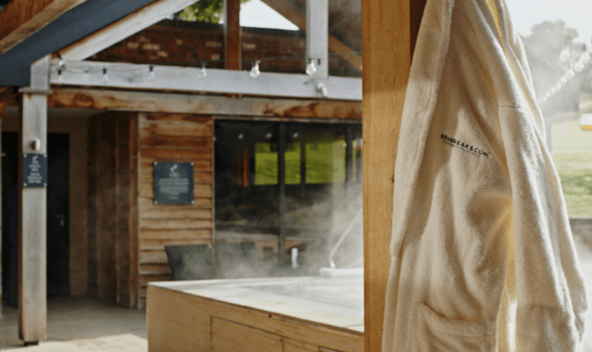 White Spabreaks.com bathrobe hanging beside a steaming outdoor hot tub, set against a rustic spa building with natural wood decor.