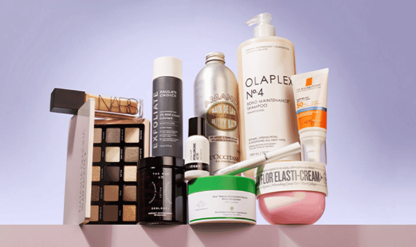 A variety of beauty and skincare products are arranged against a gradient purple background. Products include brands like Olaplex, Paula's Choice, Sol de Janeiro, L'Occitane, NARS, and others, featuring items such as shampoos, creams, eyeshadows, and serums