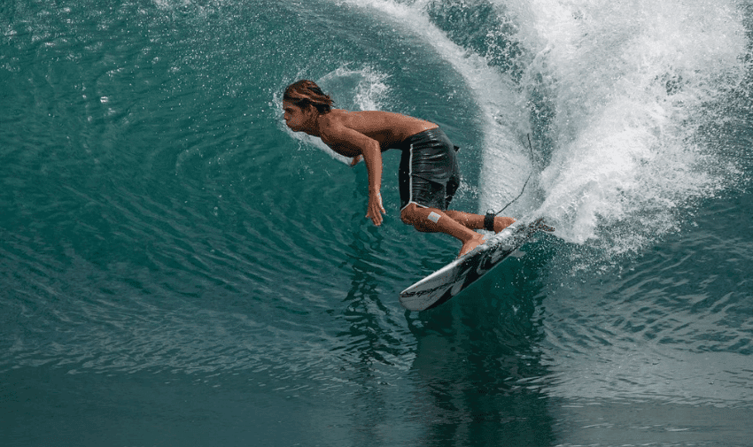  A person with long hair surfs a large, translucent blue wave. They are standing on a surfboard, wearing black shorts, and leaning forward with water spraying behind them. The wave curls dramatically, creating a dynamic and thrilling scene.