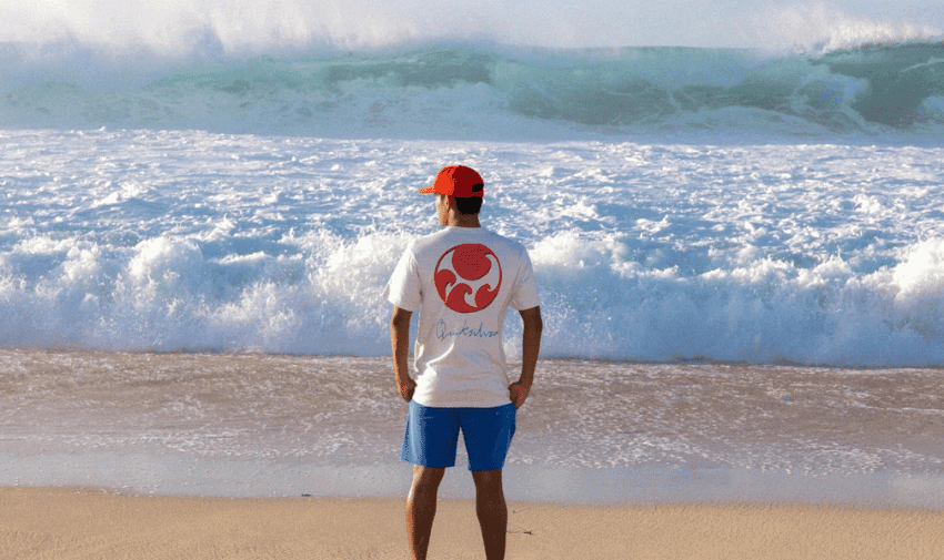  A person wearing a white shirt with a red circular design, blue shorts, and a red cap stands on a sandy beach facing the ocean. Large waves are crashing in the background.