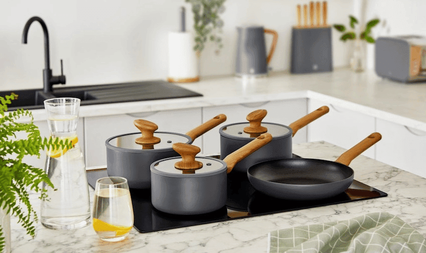  A modern kitchen features a white marble countertop with a set of four SWAN grey pots and a frying pan, all with wooden handles, placed on a black stove. There is a glass pitcher and a glass of lemon water nearby. A green checkered cloth is draped on the counter.