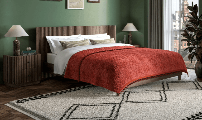 A cosy bedroom with a wooden bed frame, white sheets, and a red patterned blanket. The walls are green, and side tables hold decorative lamps. A large window with curtains offers a city view, and a beige rug with black patterns covers the floor.