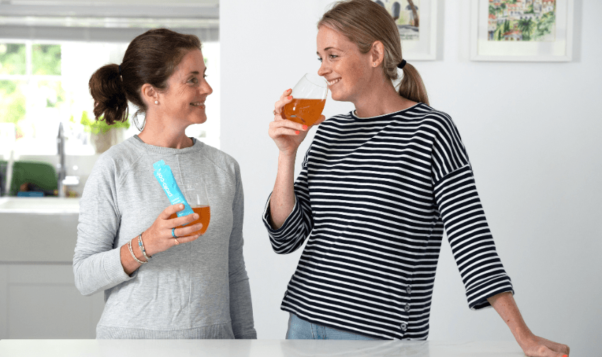 Two women in a bright kitchen enjoying Proto-col collagen drinks, one holding a sachet of the supplement. Keywords: Collagen Supplements, Beauty and Wellness, Health Support for Disabled People.