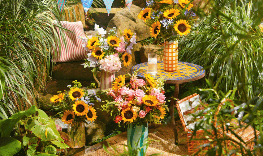 A vibrant outdoor setting with various colourful bouquets of flowers, including sunflowers and roses, placed in vases on and around a small mosaic table. Green foliage surrounds the scene, and a pink-and-white striped cushion rests among the plants.