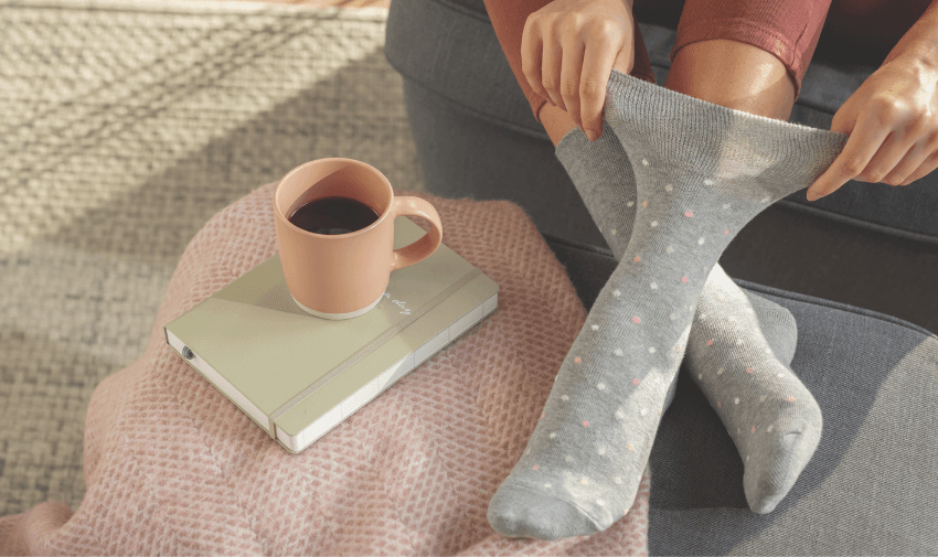 someone sat on the floor pulling their sock up, on the floor next to them, there is a mug of coffee on top of a pale green notepad.