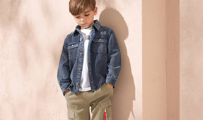 A young boy with short brown hair stands against a light peach-coloured wall. He is wearing a denim jacket over a white T-shirt and green quilted pants with pockets, hands in his pockets, looking down thoughtfully.