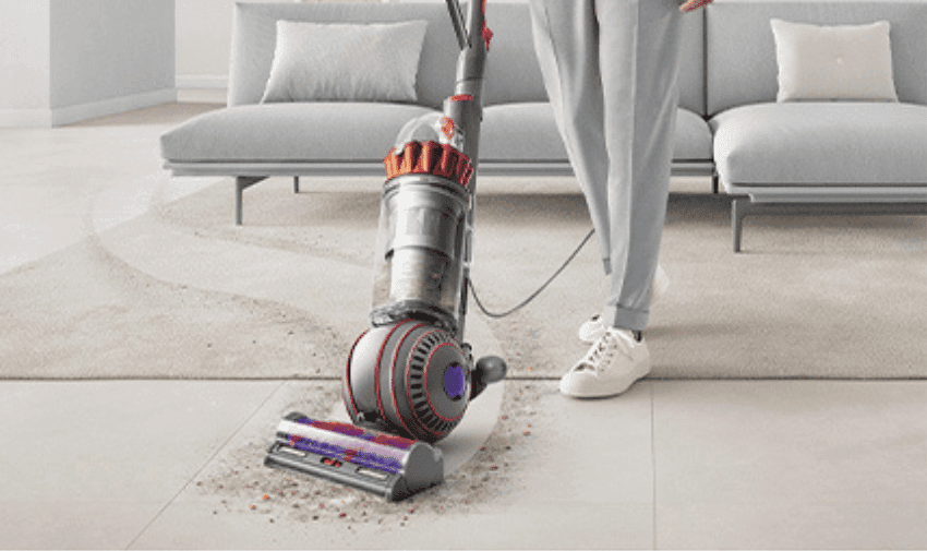 A person in grey pants and white shoes is vacuuming a light-coloured carpet in a modern living room with a Dyson. The vacuum cleaner is picking up dirt and debris from the carpet, leaving a clean path behind. A grey sofa is visible in the background.