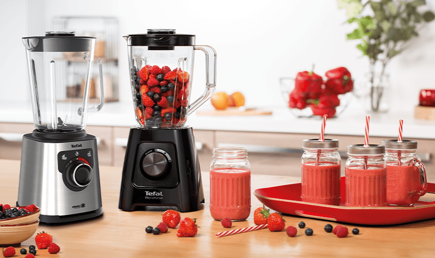 Two Tefal blenders, one blending red fruits, sit on a kitchen counter surrounded by assorted berries. On the right, two mason jar glasses filled with berry smoothies are placed on a red tray. The bright kitchen is decorated with fruits and herbs.