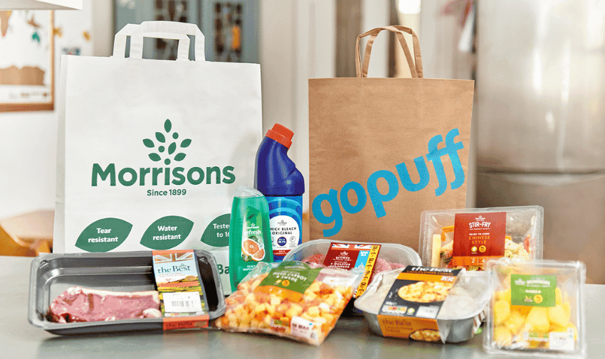 A variety of groceries are displayed on a kitchen counter, including packaged meat, fresh produce, a bottle of bleach, body wash, and ready-made meals. There are two grocery bags in the background; one is white with the Morrisons logo, and the other is brown with the GoPuff logo.