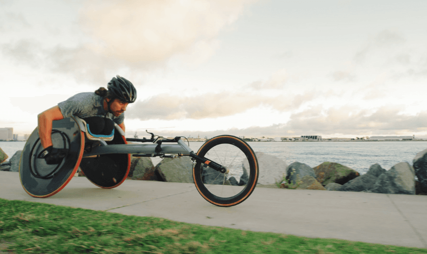 A male athlete in a racing wheelchair speeding along a scenic seaside path. Explore exclusive sports and fitness discounts for disabled people with Purpl.