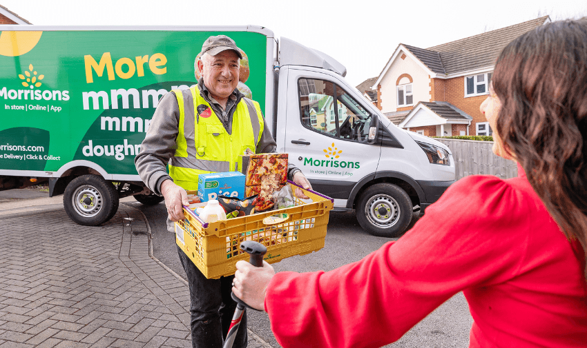 A Morrisons delivery driver hands a yellow crate of groceries to a smiling woman using walking poles, with a branded Morrisons van in the background. Shopping discounts for disabled people.