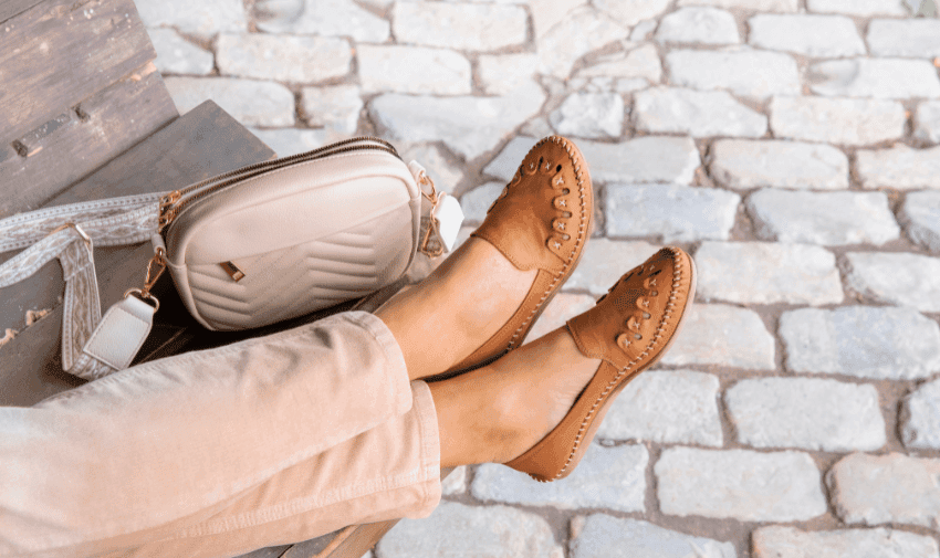 A person wearing brown leather slip on shoes rests their feet on a bench. Beside them is a beige crossbody bag with a quilted pattern. The ground is paved with large, uneven stones.