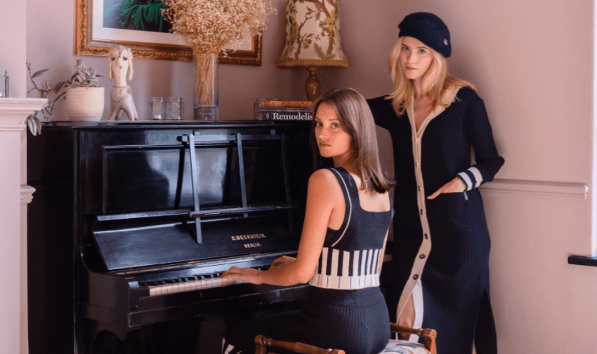 Two women in a cosy room, one plays the piano while the other leans against it, dressed in chic, retro-inspired outfits perfect for stylish occasions.