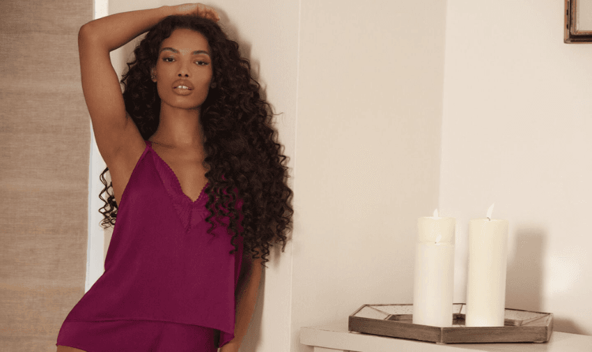 A person with long, curly hair is wearing a matching magenta top and shorts set, standing next to a small table with lit candles. The setting is indoors, with neutral-colored walls.