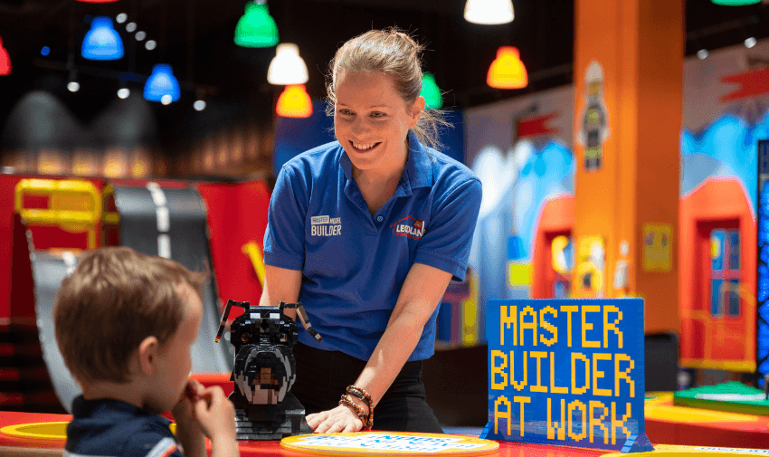 A smiling Legoland Discovery Centre employee, dressed in a blue uniform, helps a young child with a LEGO construction project. A "Master Builder at Work" sign adds to the excitement of the hands-on experience. Disabled travel discounts UK, inclusive attractions, and accessibility for families.