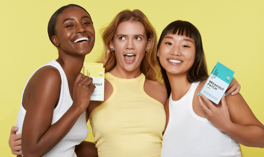 Three women holding skincare products against a yellow background, celebrating diversity and clear skin. Ideal for skincare product discounts for disabled cardholders.