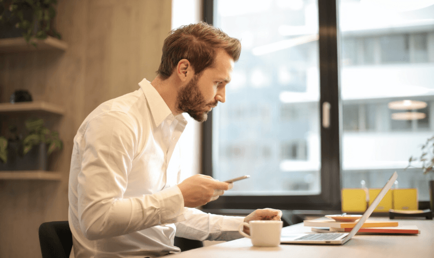 A man at his desk intently reviewing details on his laptop and smartphone, representing staying on top of finances and boosting credit scores with Purpl and Credit Ladder.