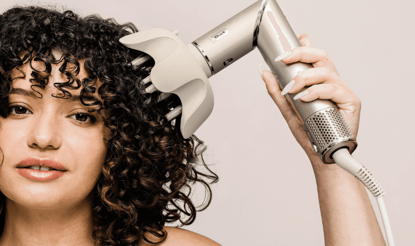 A person with curly hair uses a diffuser attachment on a hairdryer. The diffuser is silver and the person is gently holding it against their hair, which falls in defined curls. The background is a soft, neutral colour