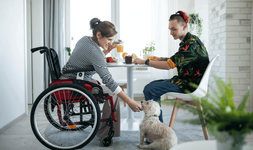 A woman in a wheelchair and a man are sitting at a dining table, enjoying a meal together. The woman, who looks happy, is wearing a striped shirt and petting a small dog. The man, dressed in a colourful shirt, appears to be mid-conversation. The setting is a bright, modern room reflecting inclusivity and warmth.