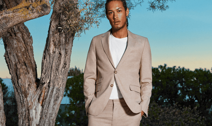 A man with long hair stands outdoors next to a tree. He is wearing a beige suit with a white shirt underneath. The background features green foliage and a serene sky with hints of blue and orange.