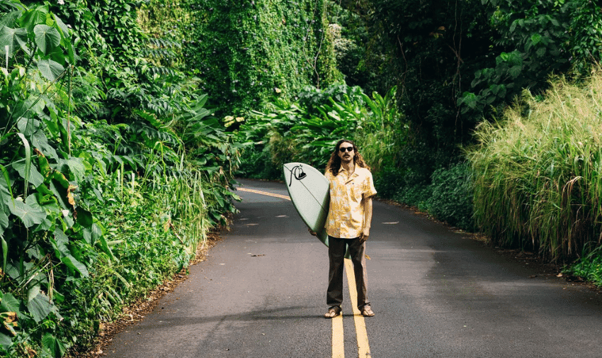 A person with long hair, wearing a yellow floral shirt, dark pants, and sandals, stands in the middle of a lush, green, forested road holding a surfboard. The road is lined with dense tropical vegetation.