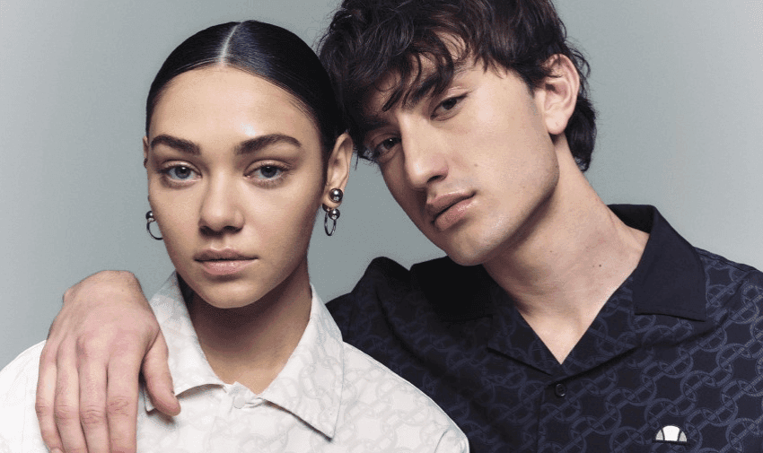  A young woman with dark hair tied back and a young man with tousled dark hair pose together. The woman wears a white shirt and hoop earrings, and the man wears a dark shirt, resting his arm on the woman's shoulder. Both have neutral expressions, looking at the camera.