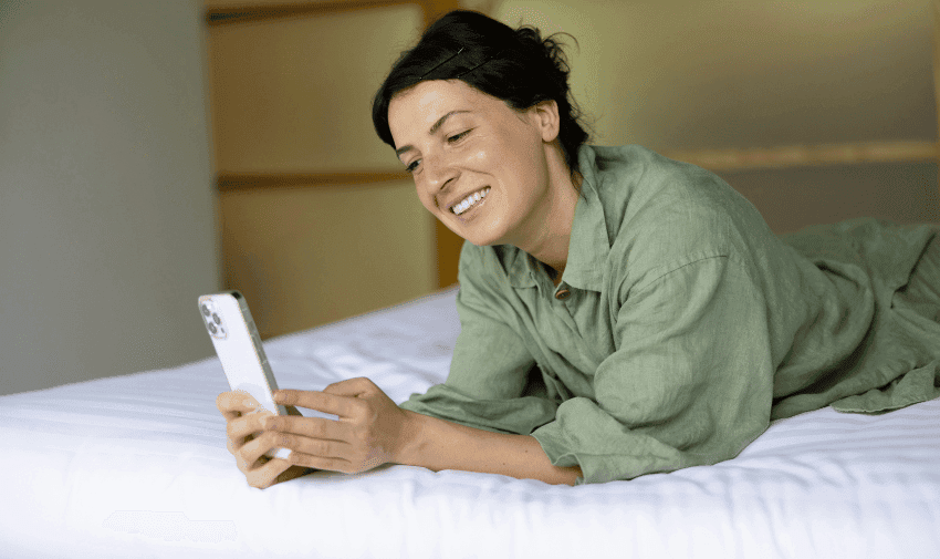 A person with dark hair, wearing a green shirt, is lying on a bed and smiling while looking at a smartphone. The background is neutral with soft lighting.