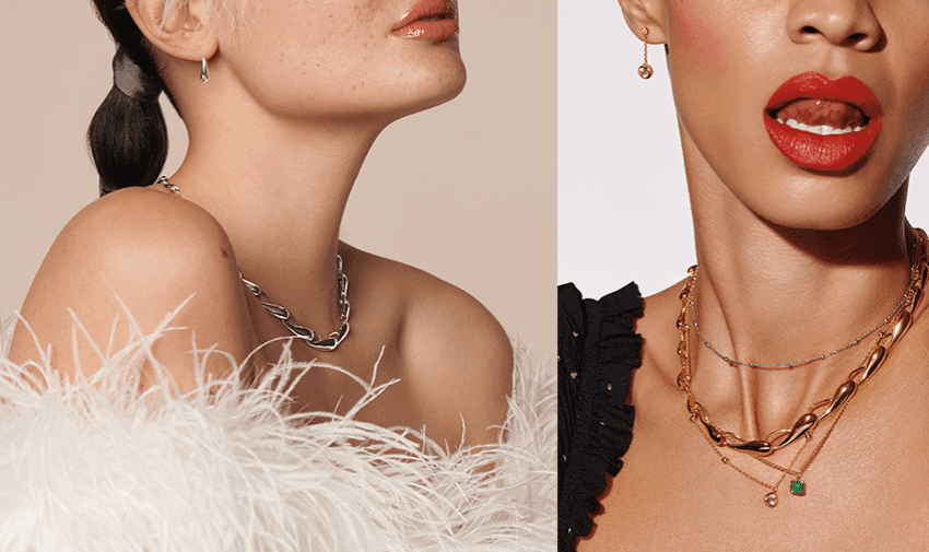 Close-up of two women wearing jewellery. The left woman has a silver chain necklace, small hoop earrings, and a white feathered garment. The right woman has bold red lipstick, gold layered necklaces, one with a small pendant, and a black ruffled top.